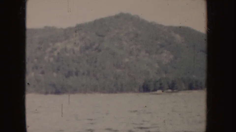 1955 Idaho 8mm frame with a soft-focus wooded hill, shoreline trees, and rippling water in the foreground.