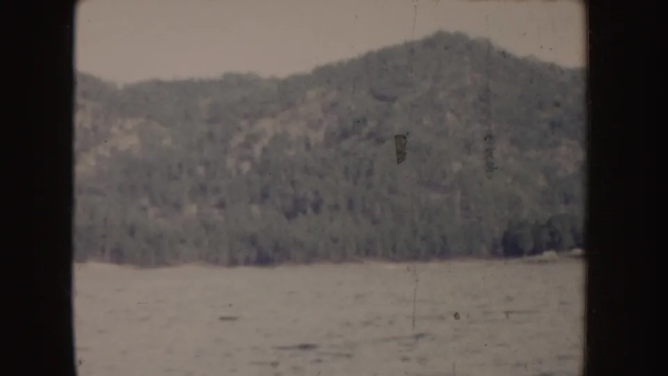 1955 Idaho 8mm frame showing a wooded hillside beyond blue rippled water, seen through dark film-gate edges.