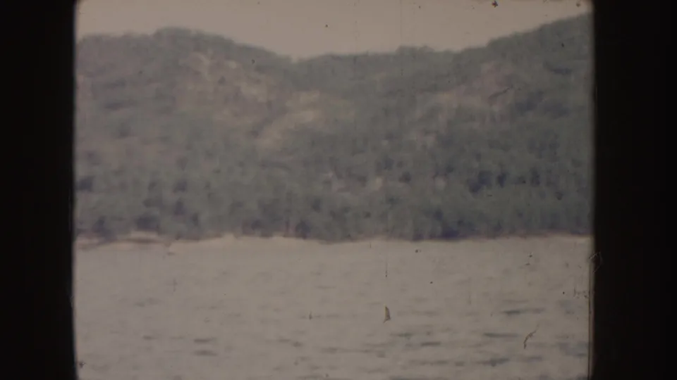 1955 Idaho home-movie view of a forested slope and shoreline across calm blue water, with visible 8mm gate framing.