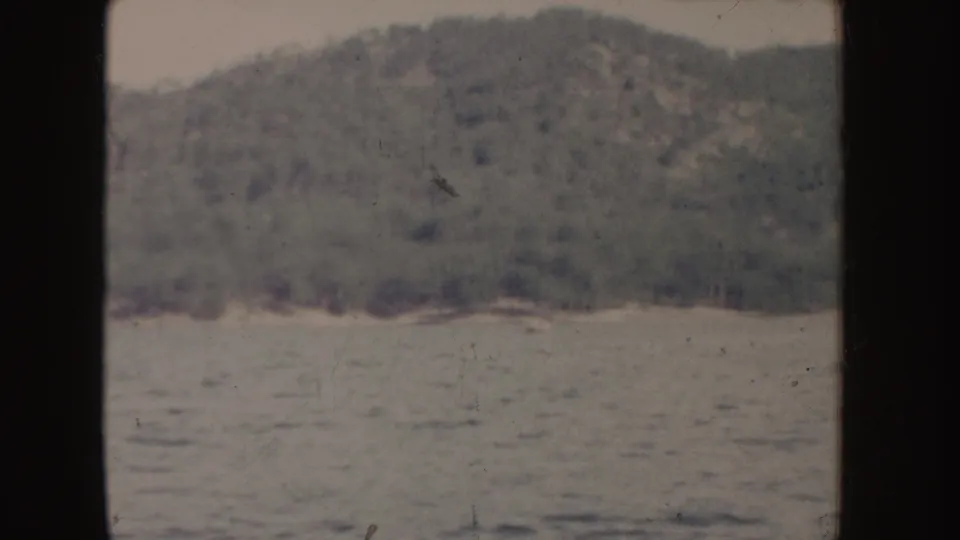 1955 Idaho 8mm still with a rounded tree-covered hill, distant shoreline, and rippled water under a pale sky.