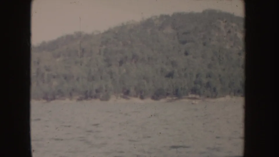 1955 Idaho 8mm frame showing a forested hillside above blue water, with dark vertical gate borders and faded color.