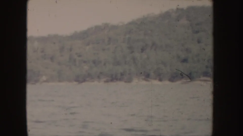 1955 Idaho 8mm frame looking across lake water toward a wooded hillside and distant shore.