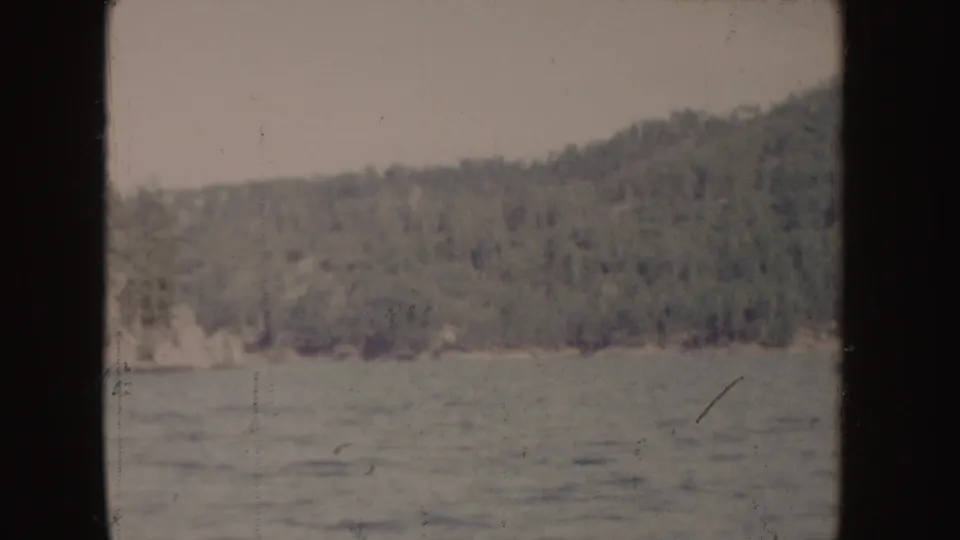 1955 Idaho home-movie frame of a wooded lake shore under muted light, filmed from a moving boat.