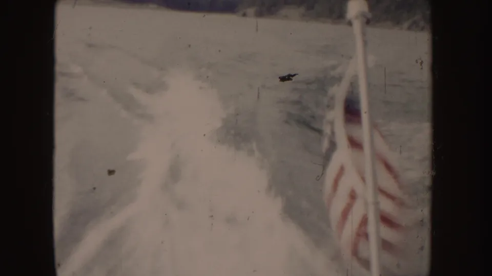1955 Idaho 8mm frame showing a small American flag on a boat with wake and wooded shoreline behind it.