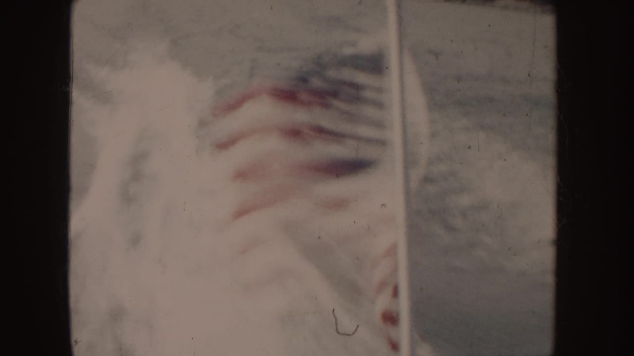 1955 Idaho 8mm frame showing a U.S. flag partly obscured by boat framing, with water and a distant shore in soft focus.