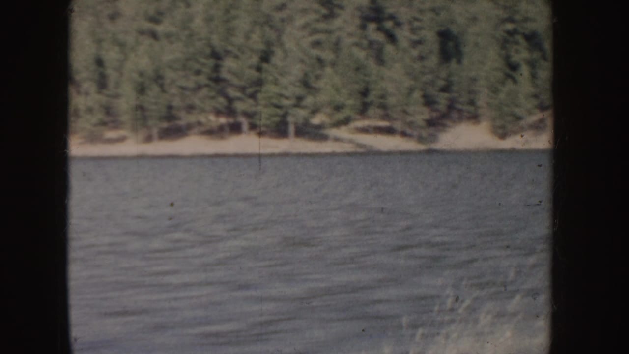 Calm lake water below a forested shoreline, framed by dark vertical film edges.