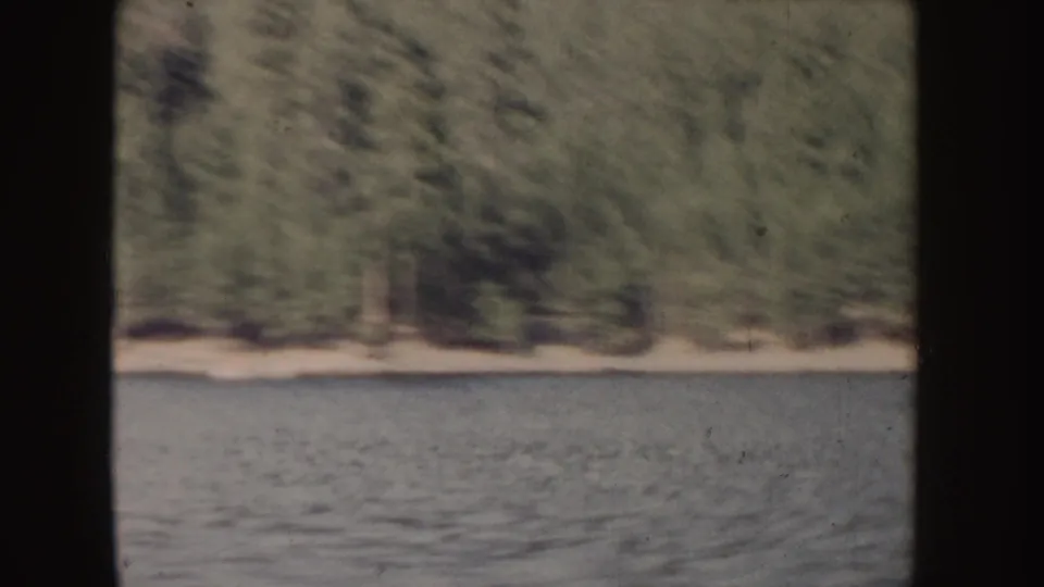 1955 Idaho 8mm frame showing a wooded lakeshore beyond rippling water, with motion blur and aged film texture.
