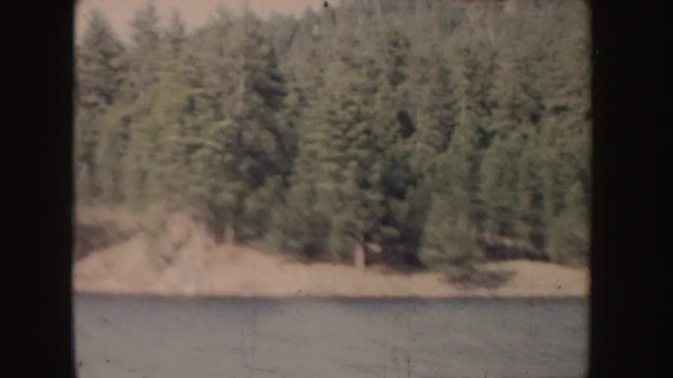 1955 Idaho lake view with an evergreen-covered shoreline or point meeting the water, seen in soft color-faded 8mm texture.