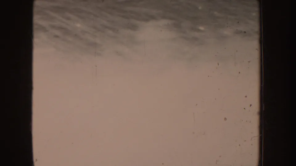 Pale 8mm frame from the lake sequence, with faint texture, dust marks, and dark gate edges but little distinct scenery.