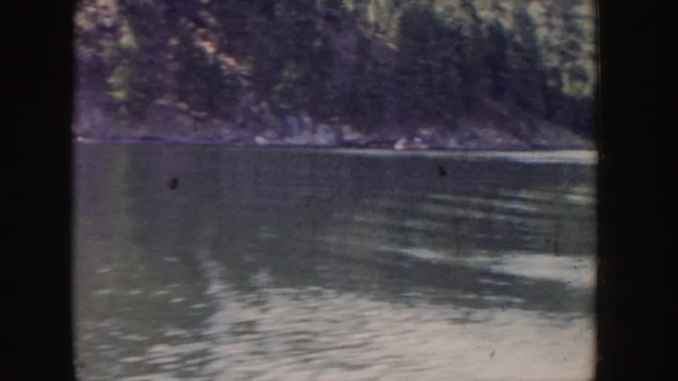 1955 Idaho lake view from a moving boat, with rippled water, white spray, and a wooded shoreline under muted light.