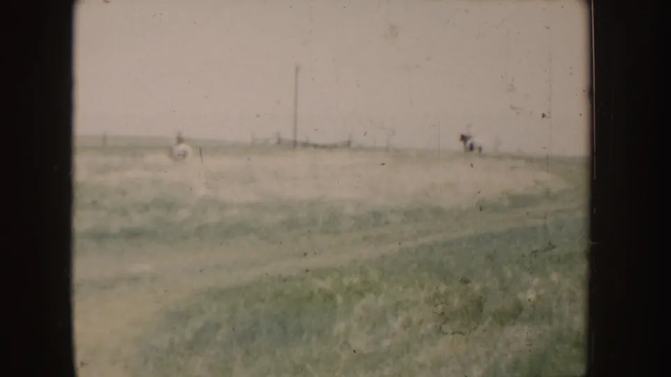 1955 Idaho home-movie frame of a broad rural field with small distant riders on horseback near the horizon.