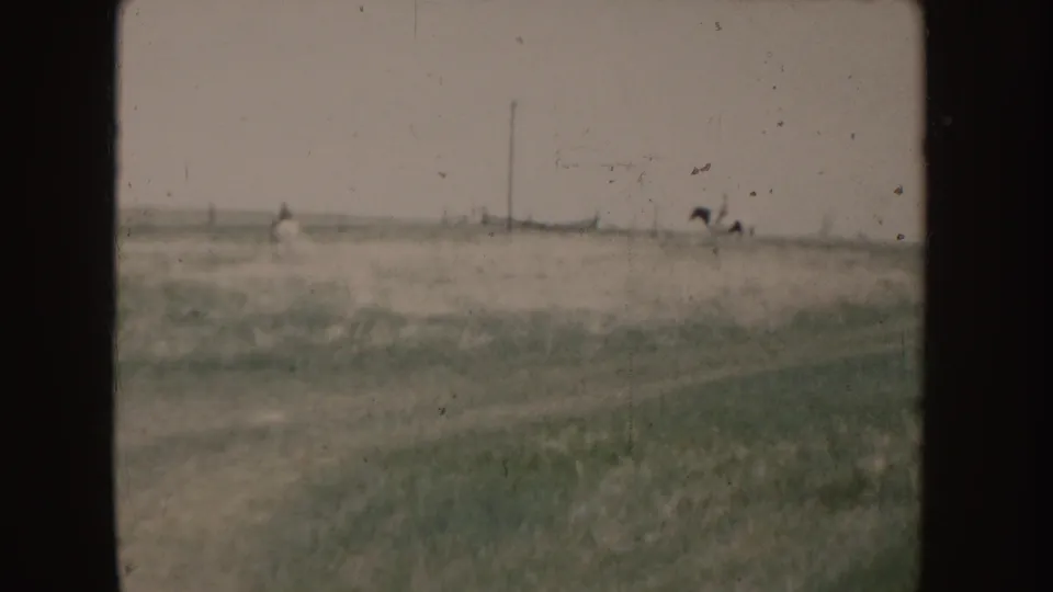 Soft 8mm frame from 1955 Idaho showing distant horseback riders moving across a wide field beneath a washed sky.