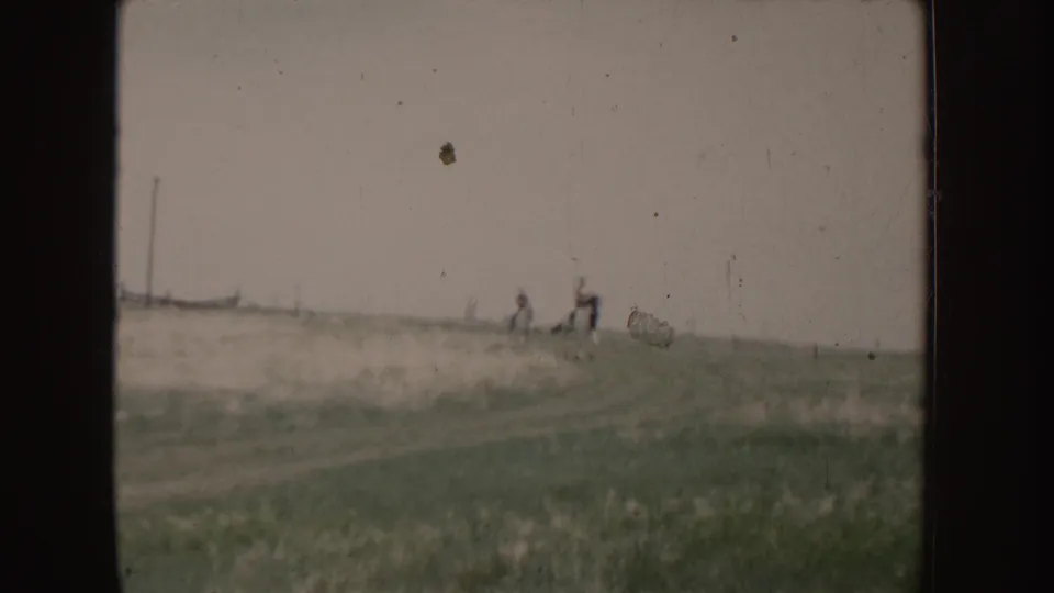 Two people ride horses across an open Idaho field in 1955 home-movie footage, seen at a distance through soft 8mm focus.