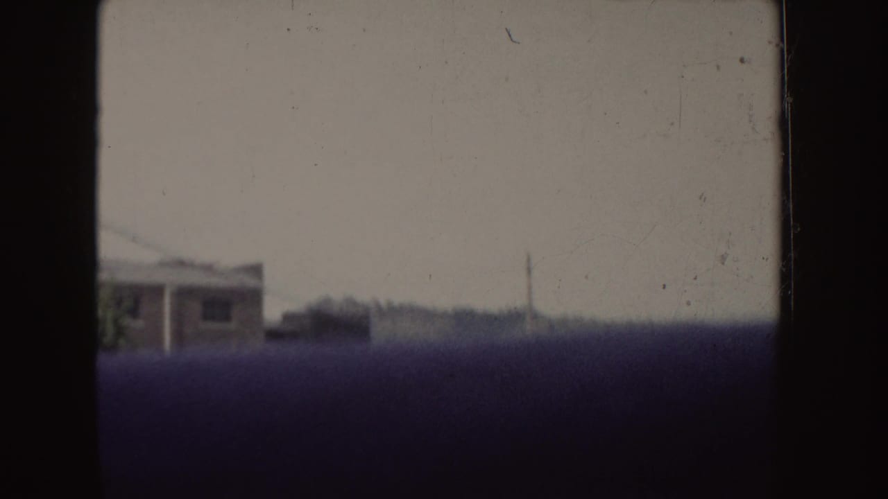 1955 Idaho 8mm frame showing a hazy moving-vehicle view past a white building, distant trees, and dark foreground obstruction.