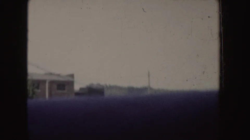 1955 Idaho 8mm frame showing a hazy moving-vehicle view past a white building, distant trees, and dark foreground obstruction.