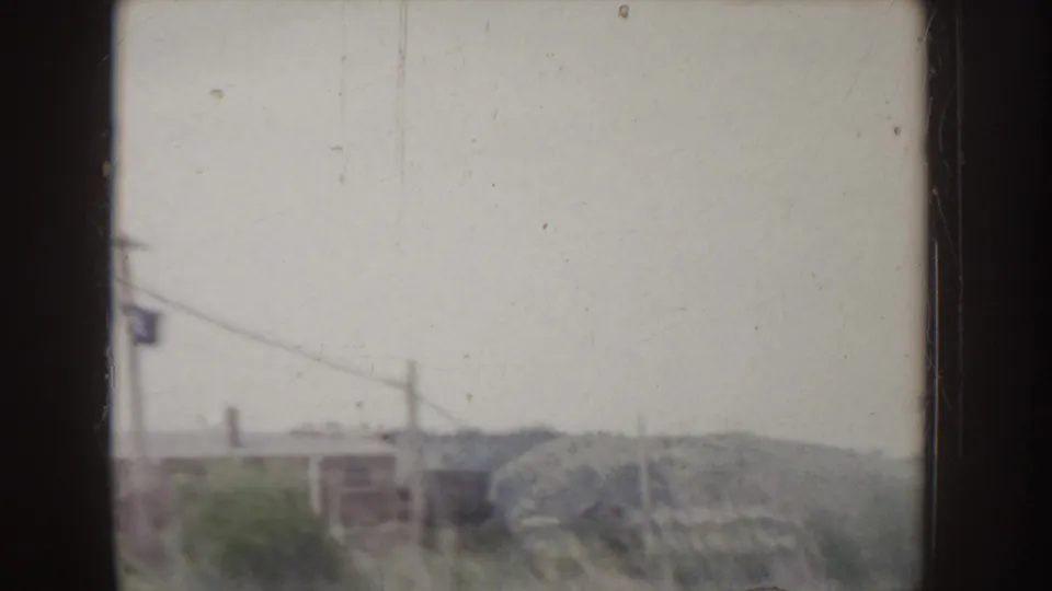 1955 Idaho home-movie frame with roadside buildings, utility lines, brushy ground, and a low hillside in soft focus.