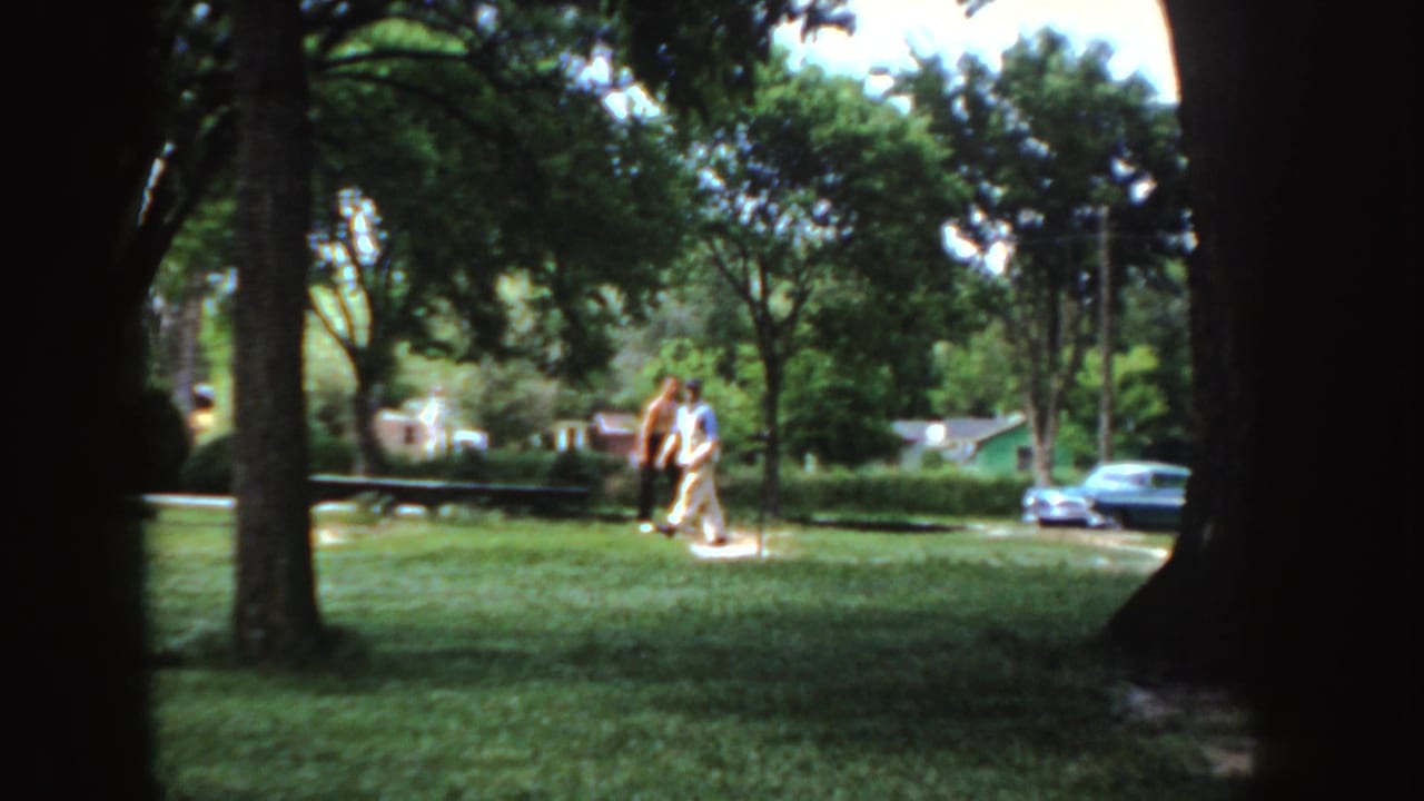 Two adults walk through a leafy Idaho park in 1955, with grass, shade trees, houses, and parked cars in the background.