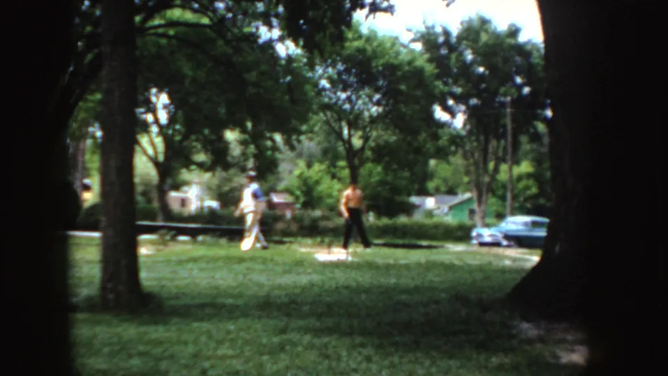 In the 1955 Idaho park scene, two adults are separated across the lawn while trees, parked cars, and houses sit behind them.
