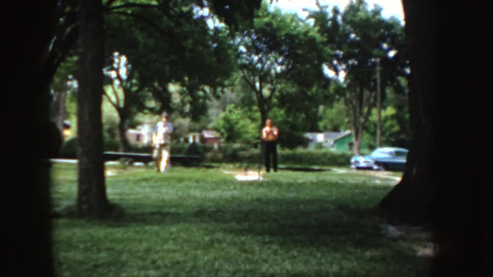 Two adults in light and dark clothing stand in a 1955 Idaho park, with a blue car, houses, and utility poles beyond the trees.