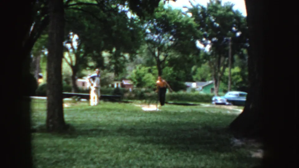 A 1955 Idaho park view shows two adults on the lawn, one with an arm raised, framed by dark tree trunks and bright foliage.