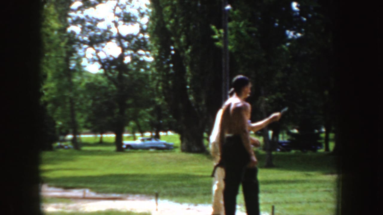 Two men stand near a horseshoe area in a sunny park with trees and parked cars behind them.