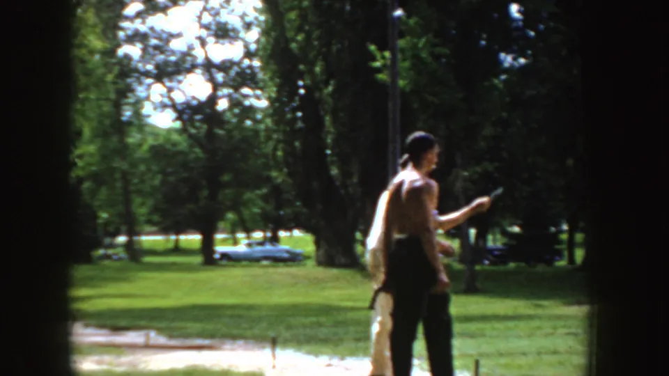 1955 Idaho 8mm frame of two adult men near a horseshoe pitch in a grassy park, with mature trees and a parked car in the background.