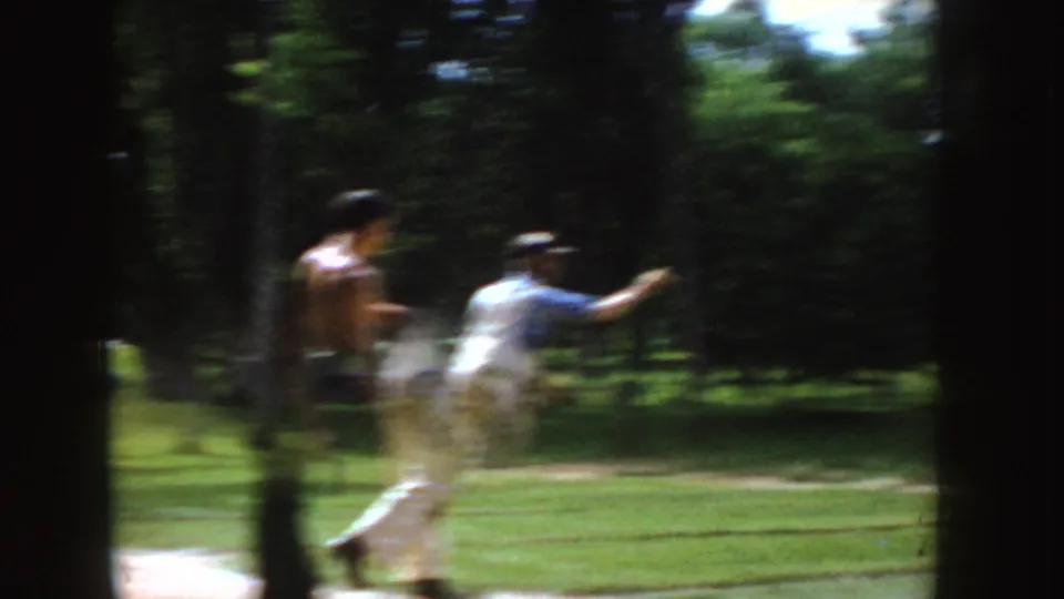 1955 Idaho home-movie frame showing a man moving through a horseshoe pitch while another player stands nearby among trees and grass.