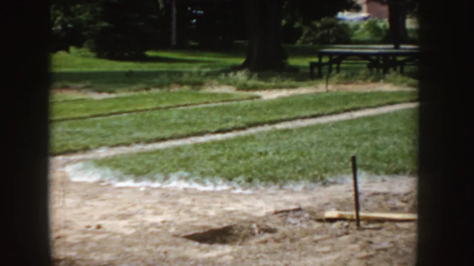 1955 Idaho 8mm frame of the horseshoe area after the throw, showing dirt lanes, grass, trees, and a picnic table in the park.