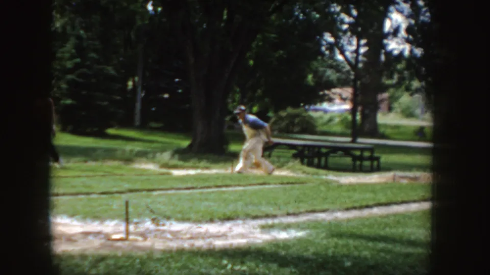 1955 Idaho home-movie frame showing a man bending near the horseshoe area, with picnic tables, grass, and mature trees behind him.