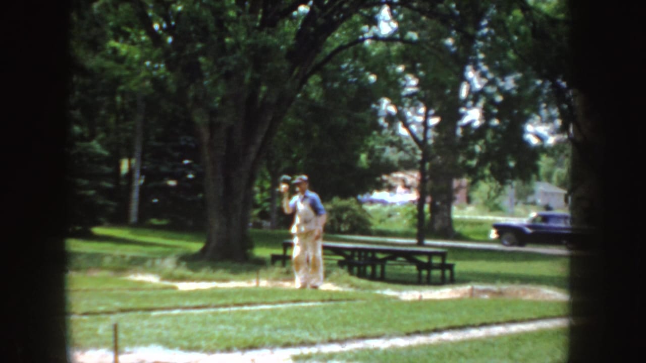 The man lifts his arm during the throw in a 1955 Idaho park, with a picnic table, large trees, and a vintage car visible.
