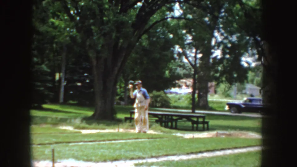 The man lifts his arm during the throw in a 1955 Idaho park, with a picnic table, large trees, and a vintage car visible.