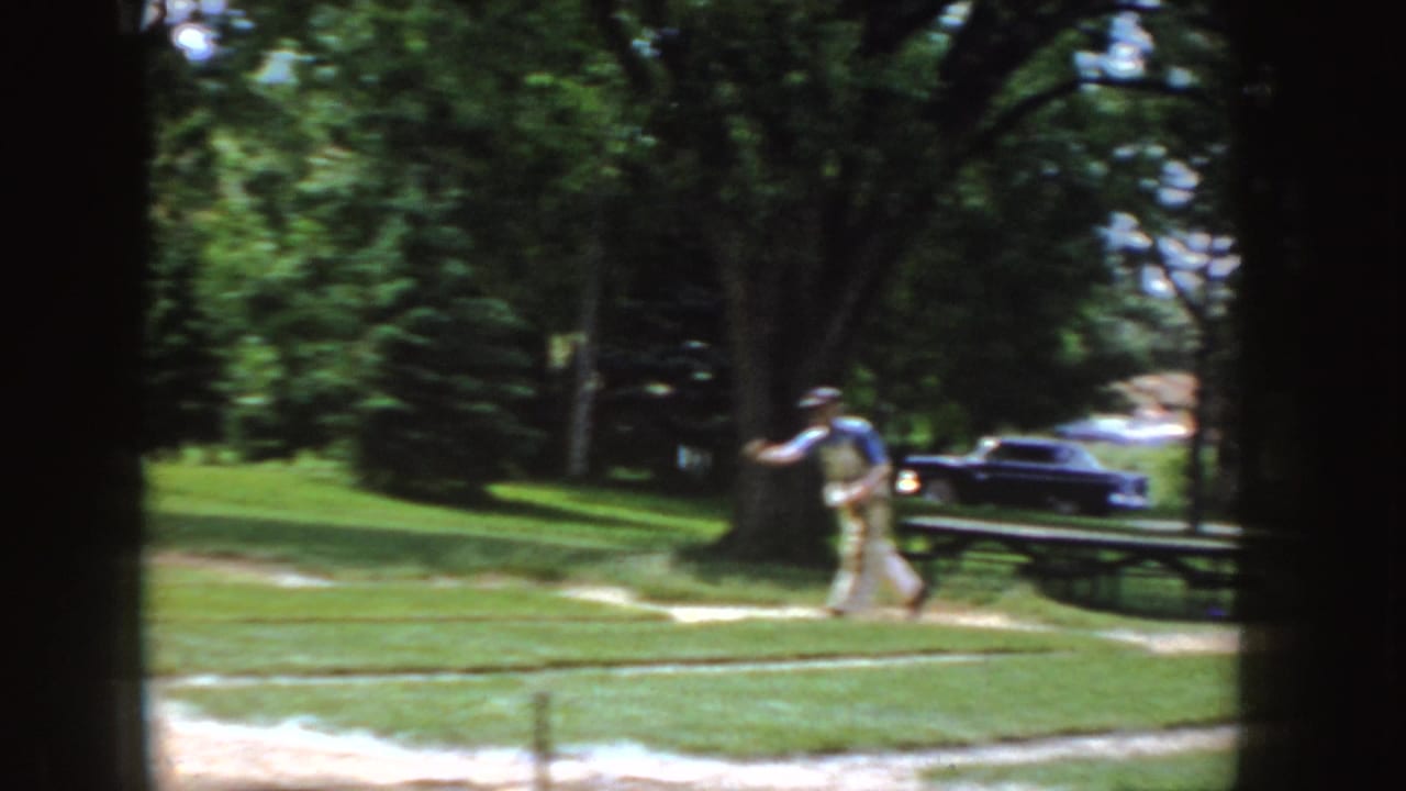 The man follows through on a pitch or throw in the grassy Idaho park, with a picnic table, mature trees, and a dark vintage car nearby.