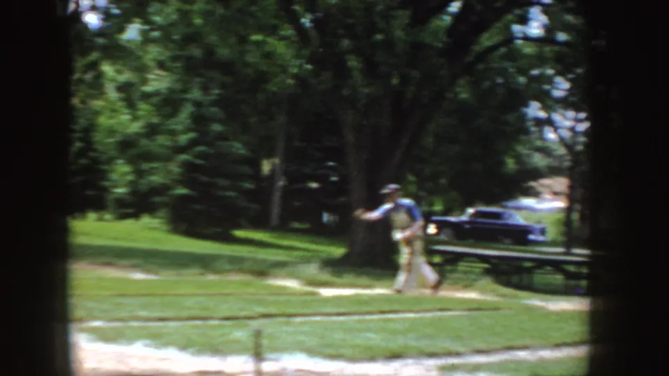 The man follows through on a pitch or throw in the grassy Idaho park, with a picnic table, mature trees, and a dark vintage car nearby.