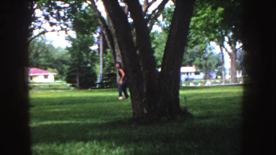 A distant person in a brown shirt and dark pants appears near a large tree in a 1955 Idaho suburban yard.