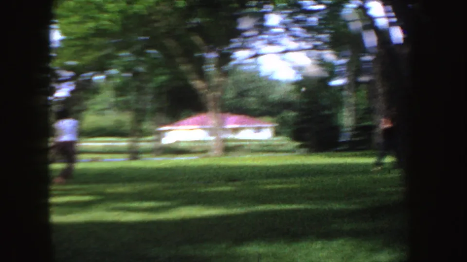 Motion-blurred 8mm frame of a person crossing a green Idaho yard as a small pale object appears near the left side.