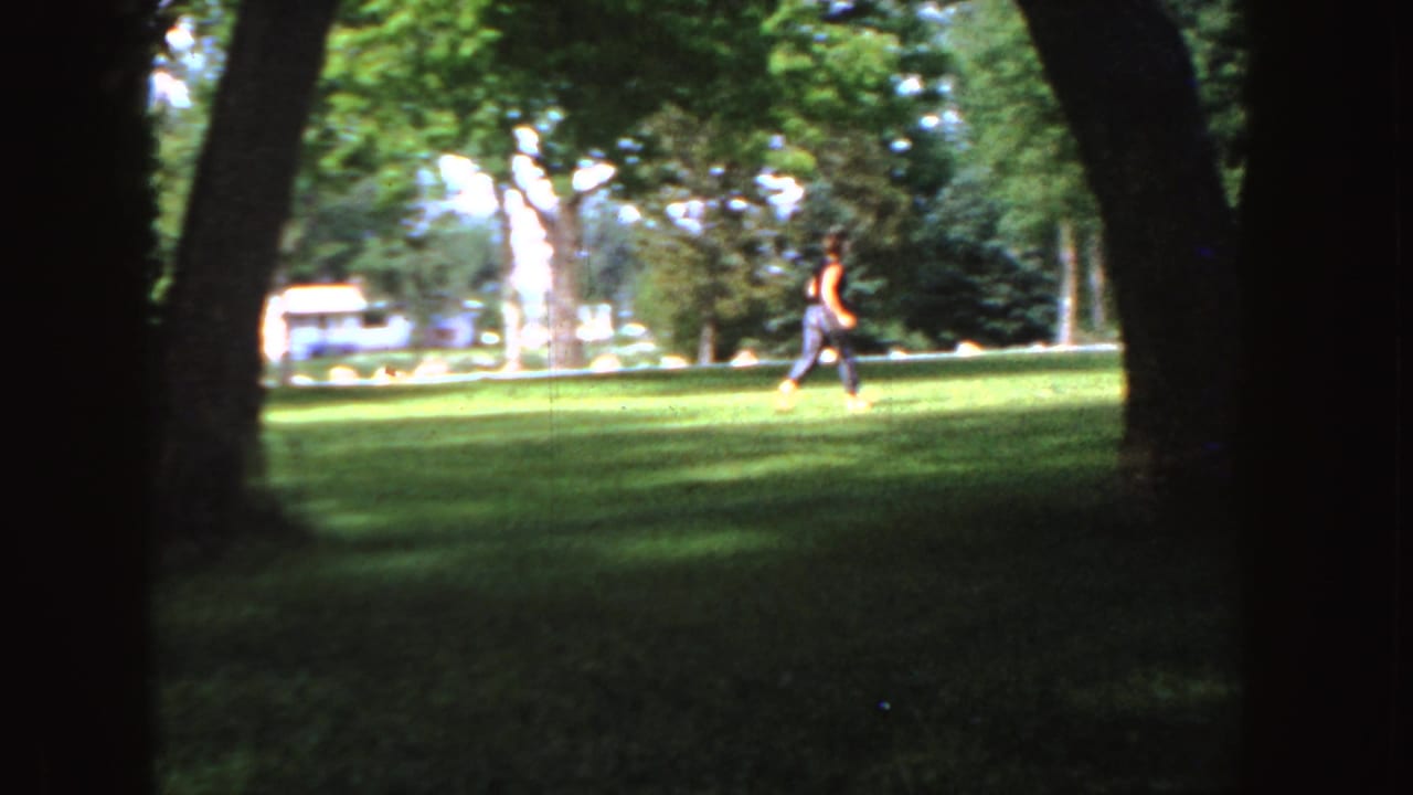 1955 Idaho 8mm frame showing a distant runner in an orange shirt crossing a sunny park lawn between large trees.