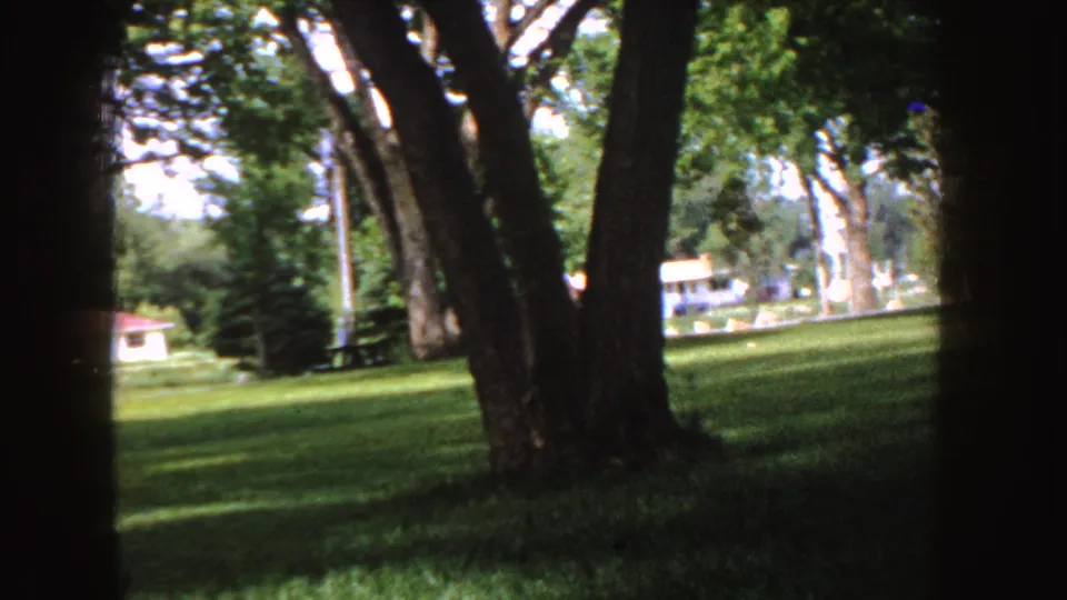 1955 Idaho 8mm frame of shaded park grass, mature trees, a picnic table, and distant buildings across the lawn.