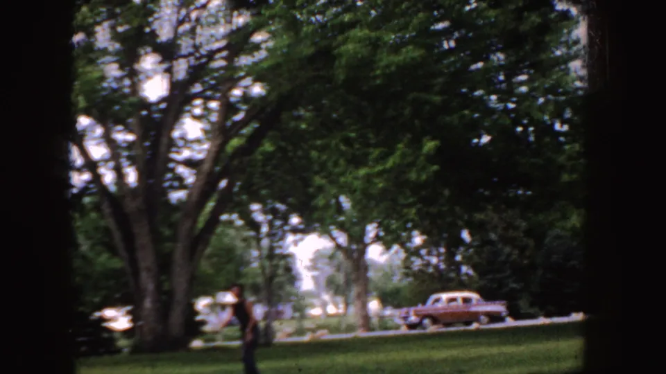 1955 Idaho park frame with a man near the foreground, heavy tree shade, and a red-and-white mid-century car parked by the road.
