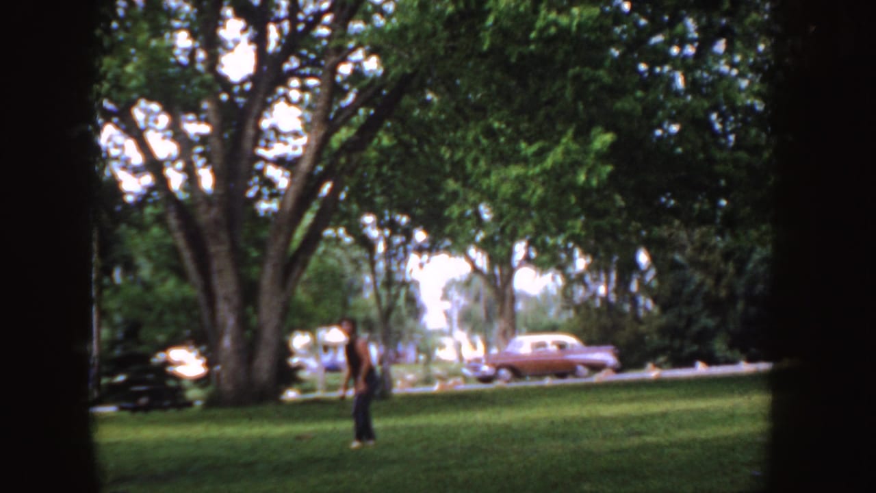 1955 Idaho 8mm frame showing a person on a grassy park lawn, large trees, and a mid-century sedan along the road.