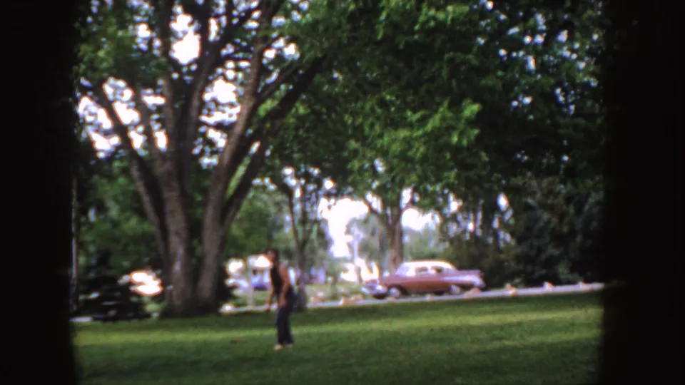 1955 Idaho 8mm frame showing a person on a grassy park lawn, large trees, and a mid-century sedan along the road.