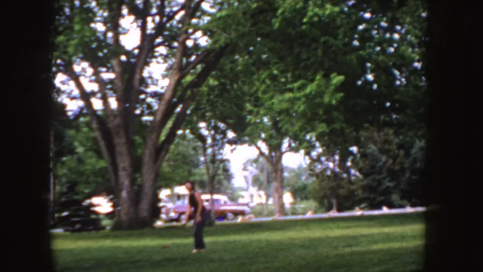 1955 Idaho home-movie frame with a person moving across a green park lawn near mature trees and a parked 1950s car.