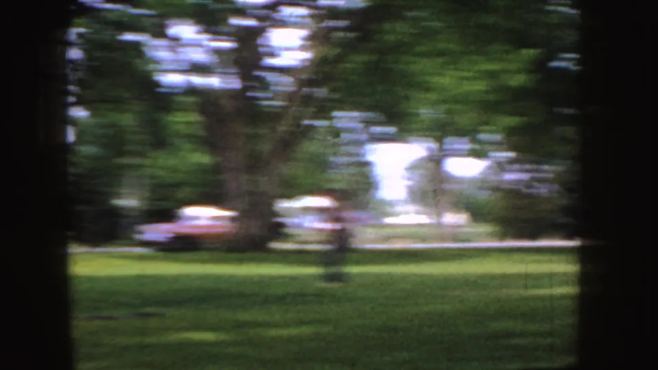 Motion-blurred 8mm frame from the Idaho park scene, with grass, trees, and a period car visible through camera movement.