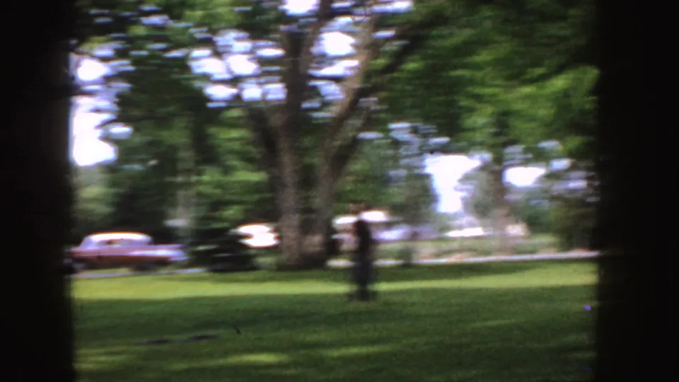 1955 Idaho 8mm frame showing a blurred figure on grass, framed by shade trees with a mid-century sedan in the background.