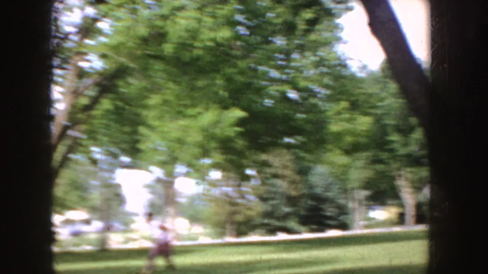 A distant person moves across a sunlit Idaho lawn in a 1955 home-movie frame, partly blurred by handheld camera motion and tree shade.
