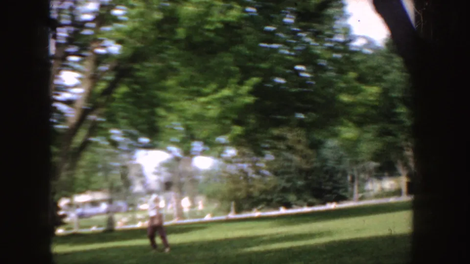 The 1955 Idaho home-movie view catches a distant person on the lawn, with mature trees, deep shade, and faint street or house details beyond.