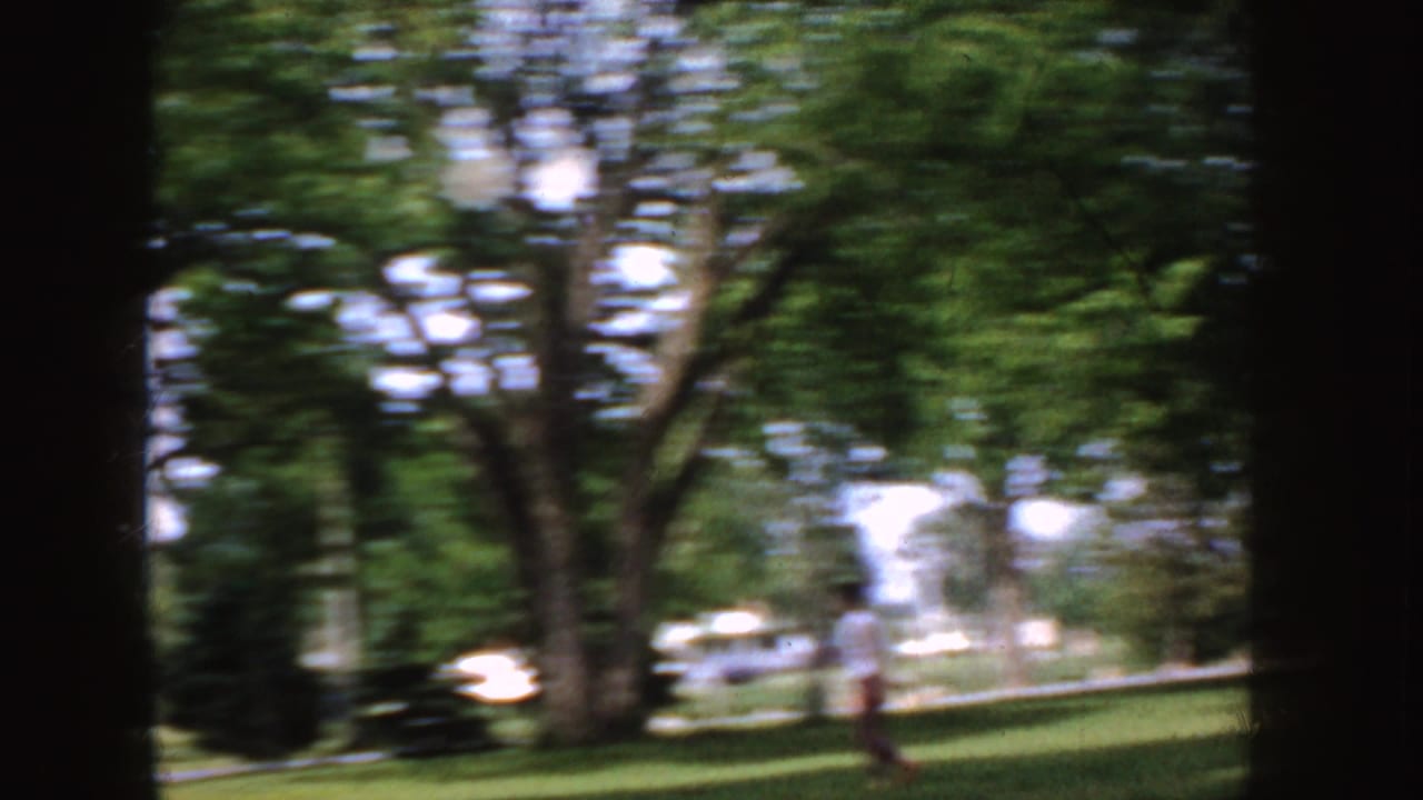 1955 Idaho 8mm frame showing a shaded suburban lawn, mature trees, a distant person, and a light-colored car near the street.