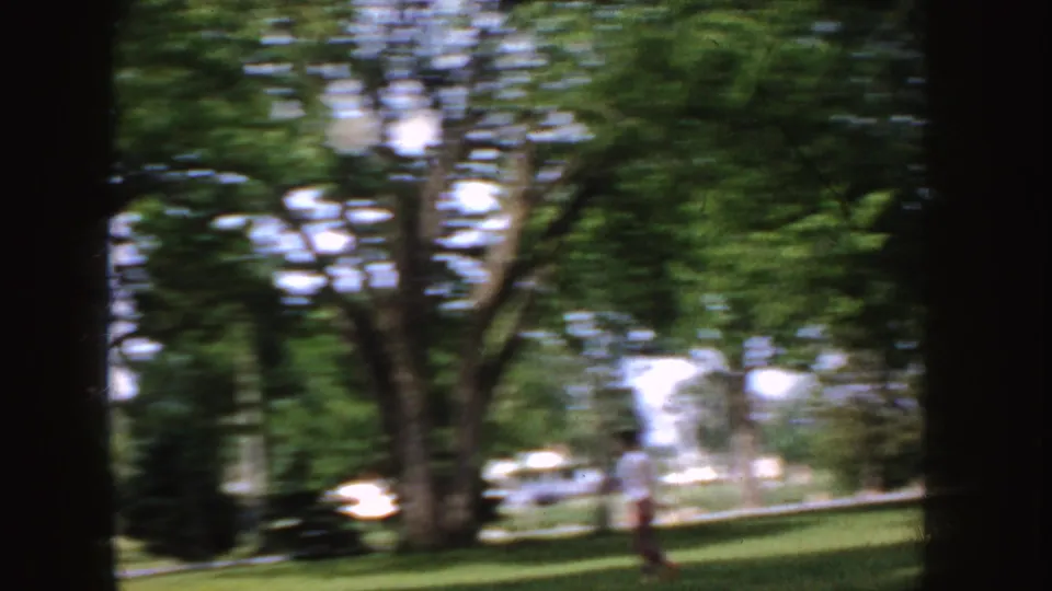 1955 Idaho 8mm frame showing a shaded suburban lawn, mature trees, a distant person, and a light-colored car near the street.