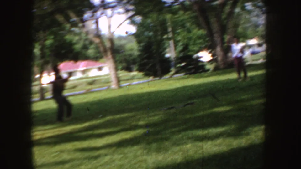 1955 Idaho 8mm frame of two distant people on a sunlit suburban lawn, with mature trees, a low house, and a parked car in the background.