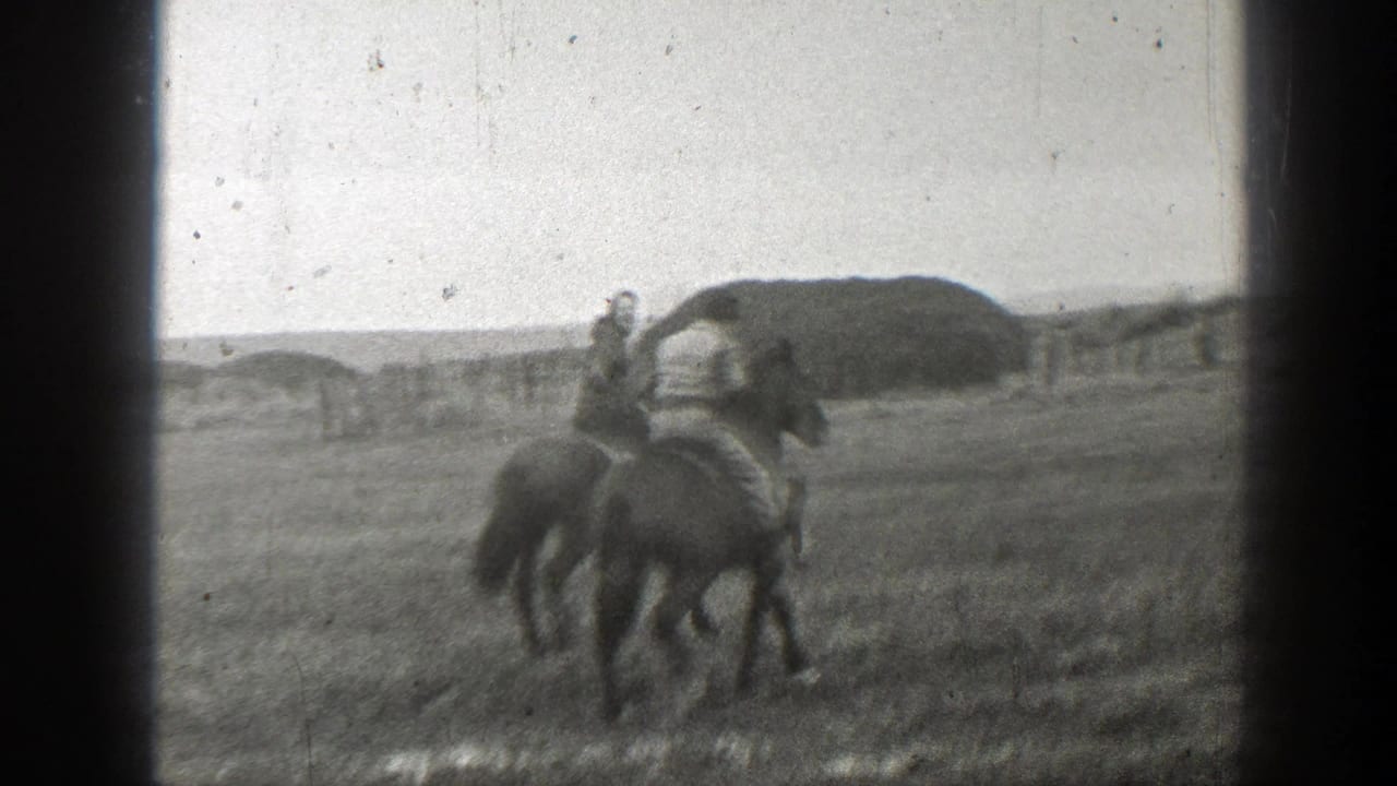In 1939 Montana footage, two people ride horses through a wet open field with a distant fence and low mound behind them.