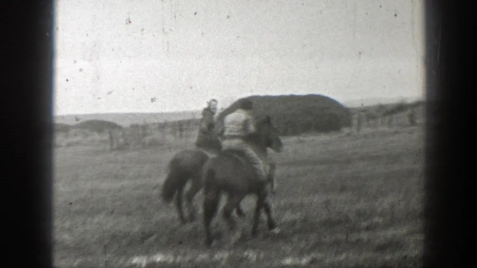 In 1939 Montana footage, two people ride horses through a wet open field with a distant fence and low mound behind them.